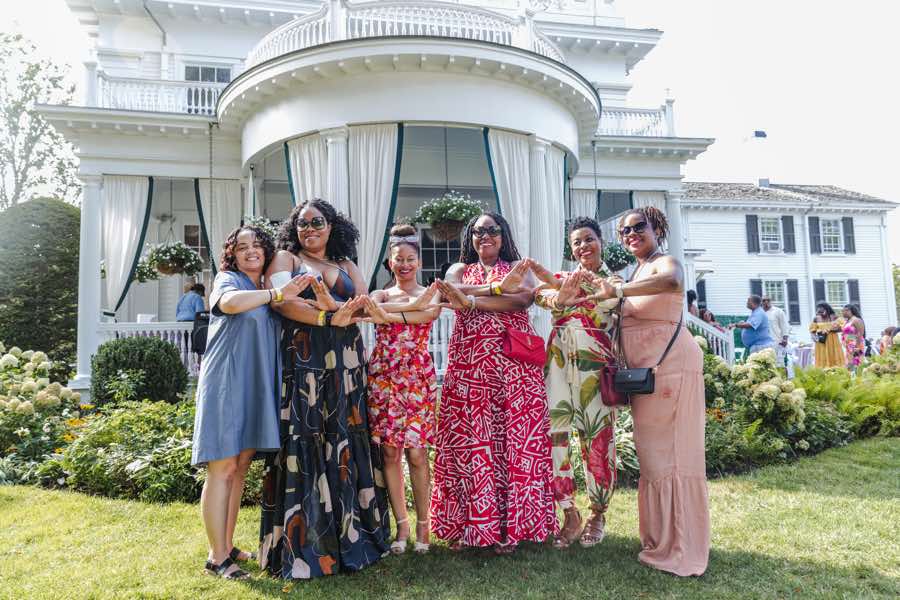 six women interlock their arms as they pose in front of a house
